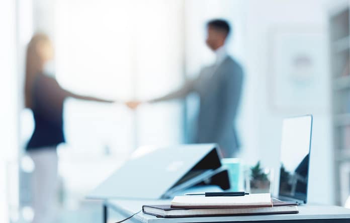 Closeup shot of a notebook and laptop on a desk in an office with two businesspeople shaking hands in the background