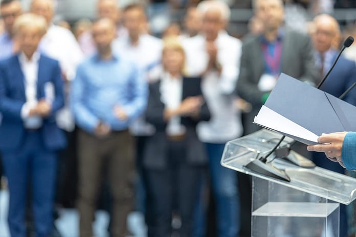 Public speaker giving presentation at business or political conference with blurred audience in background, showing leadership, communication, teamwork, and professional public speaking concept