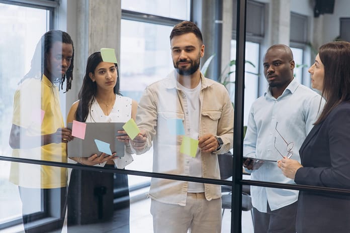 Business professionals collaborating, sharing ideas on sticky notes on a glass board, working together on a project