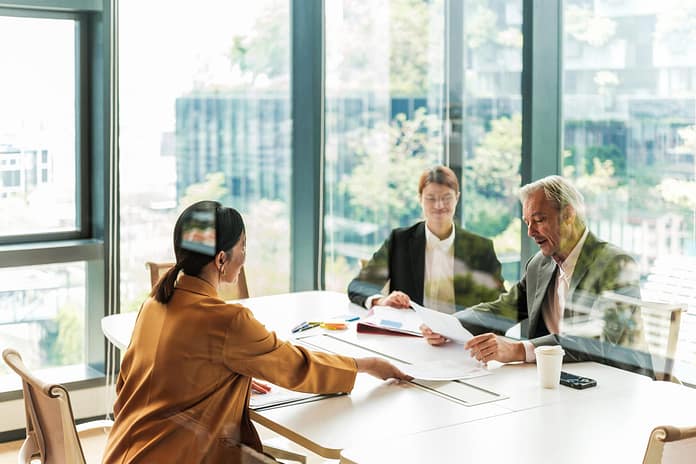 Colleagues in a bright modern office review documents around a conference table with city views.